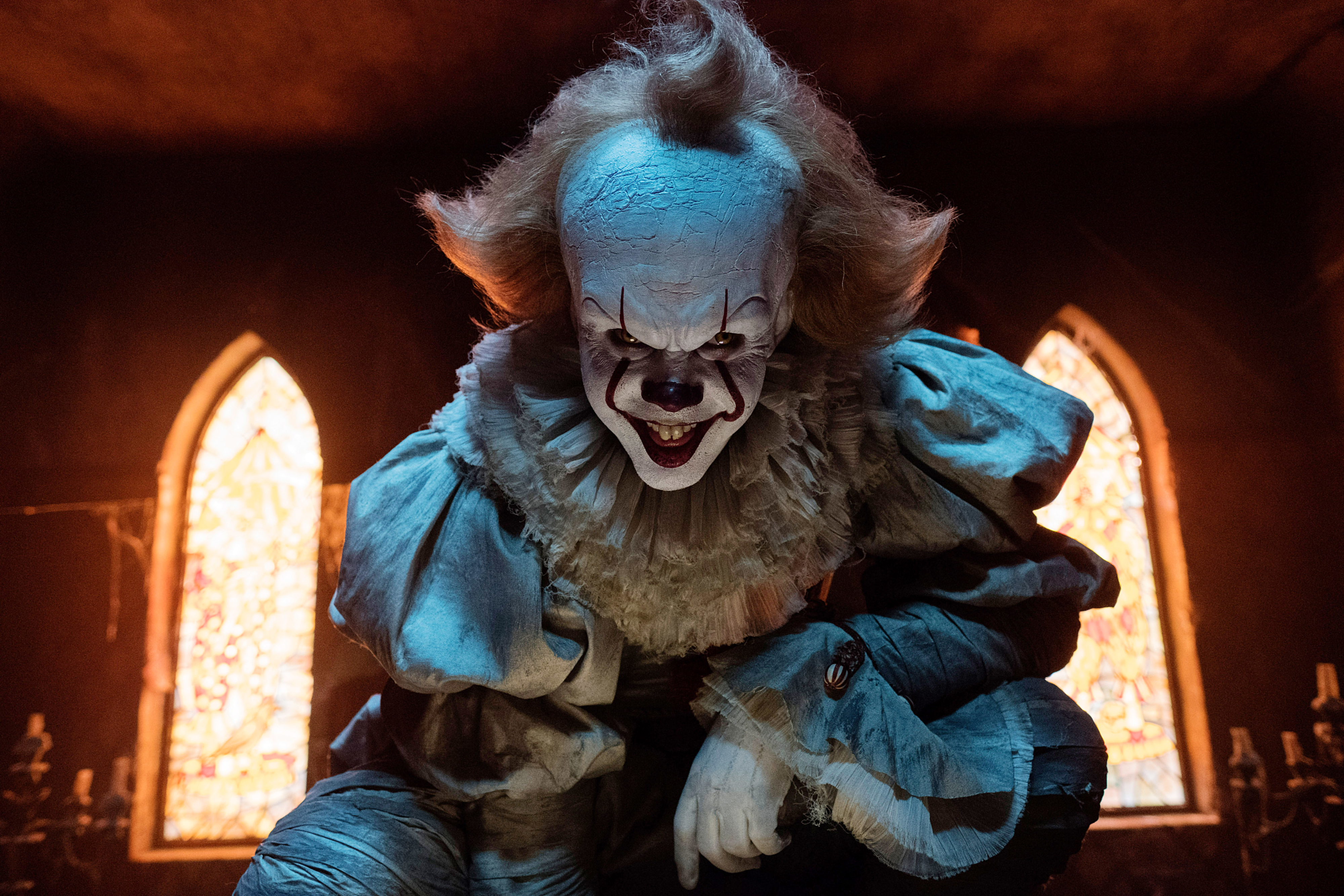 Pennywise the Dancing Clown from &quot;IT&quot; is crouched in a dimly lit room with stained glass windows in the background, wearing a vintage clown costume with eerie makeup