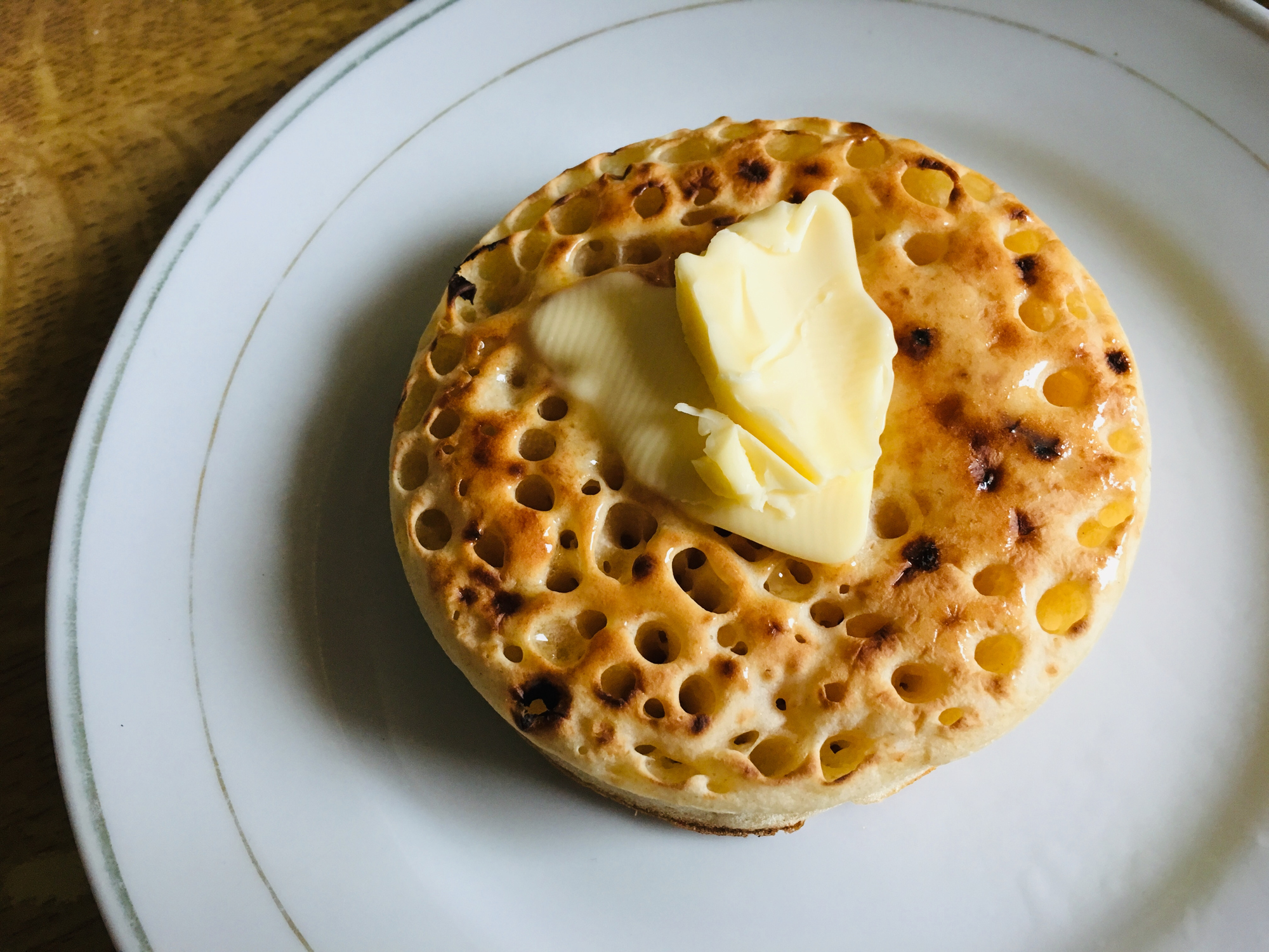 A crumpet with a dollop of butter on top sits on a white plate
