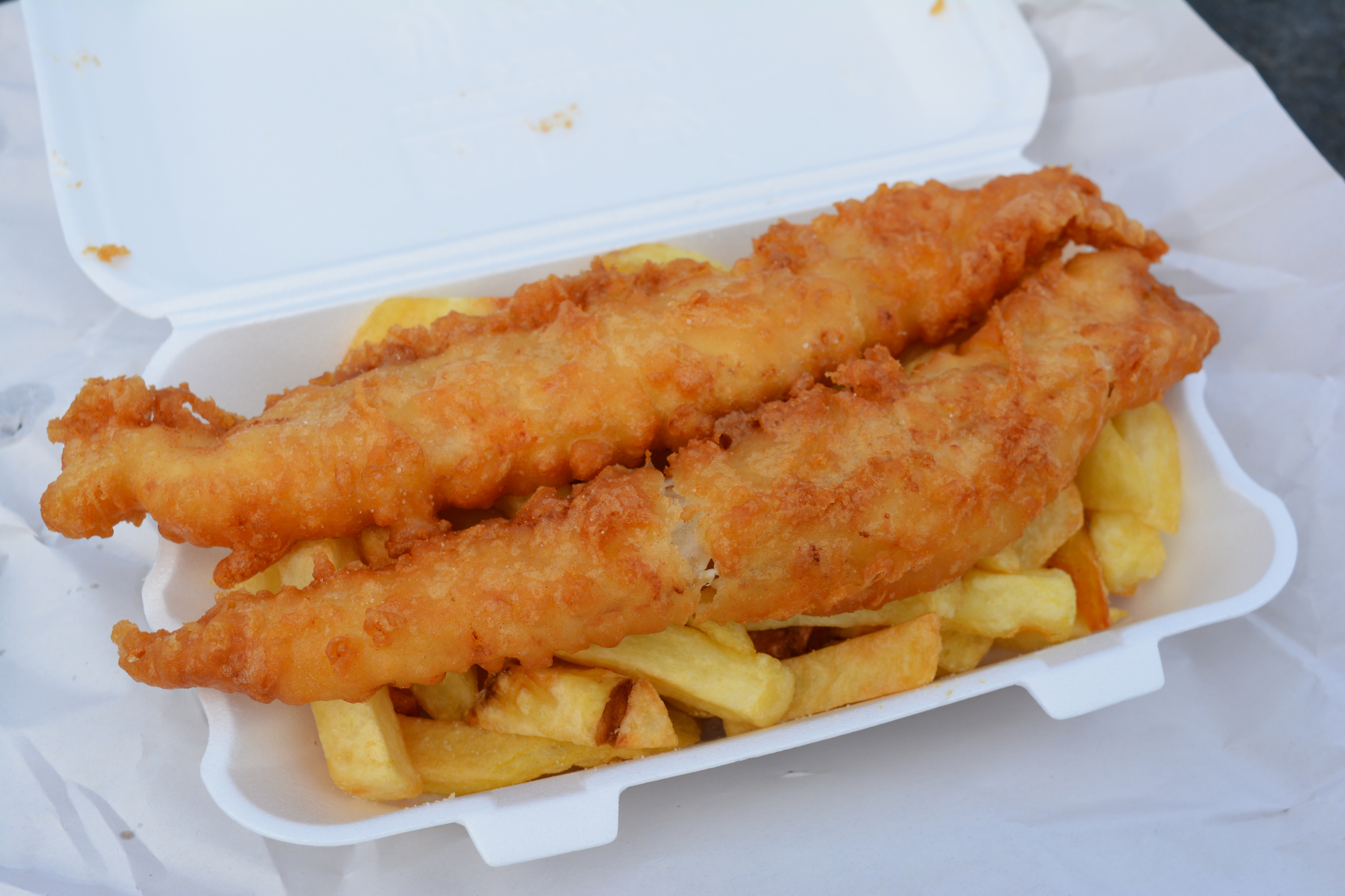 Fish and chips meal in a takeaway container featuring two pieces of fried fish on a bed of fries