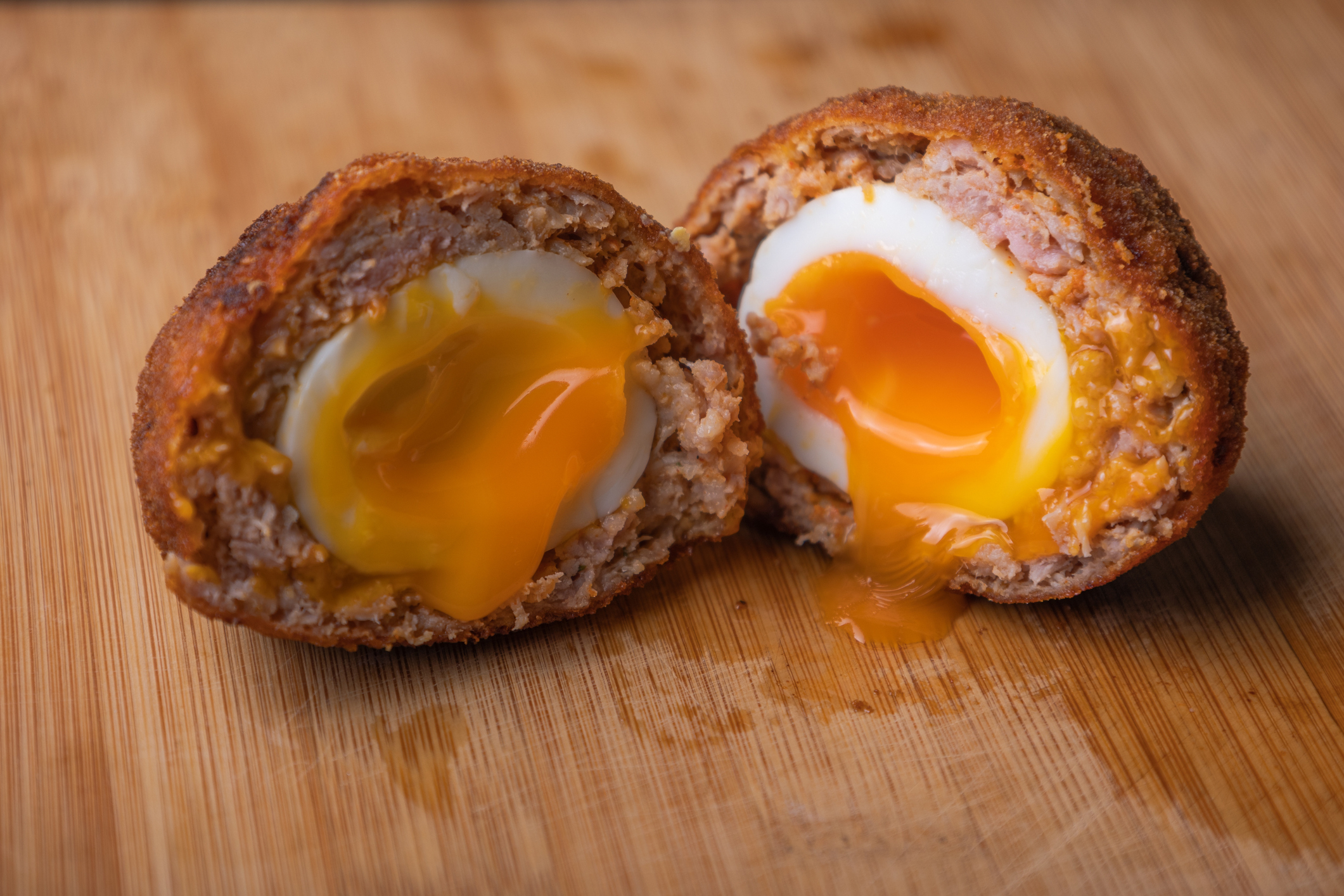 Close-up of a halved Scotch egg, with a soft-boiled egg yolk spilling out of the breaded sausage wrapping onto a wooden cutting board