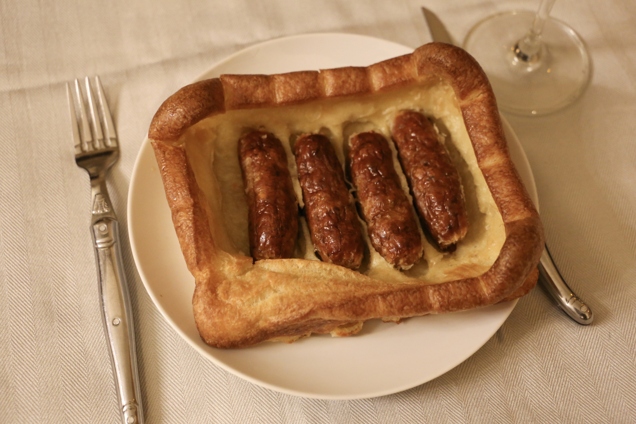 A plate with a dish of four sausages baked in a Yorkshire pudding. A knife and fork are placed beside the plate