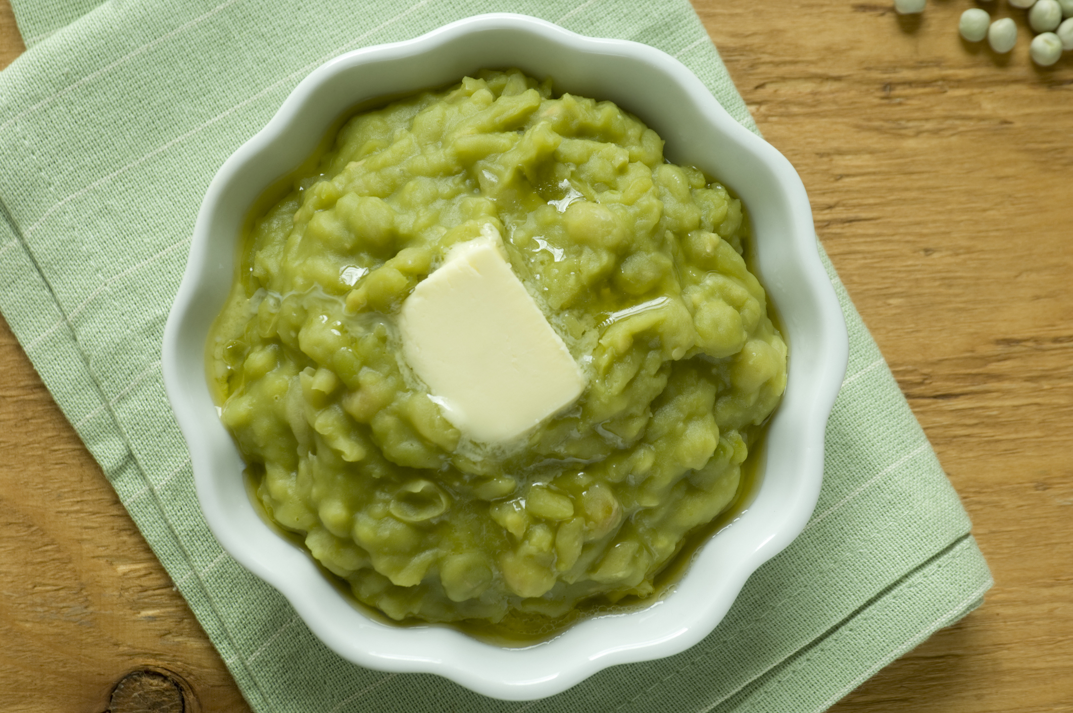 A bowl of mashed green peas with a pat of butter on top, placed on a green fabric napkin on a wooden surface