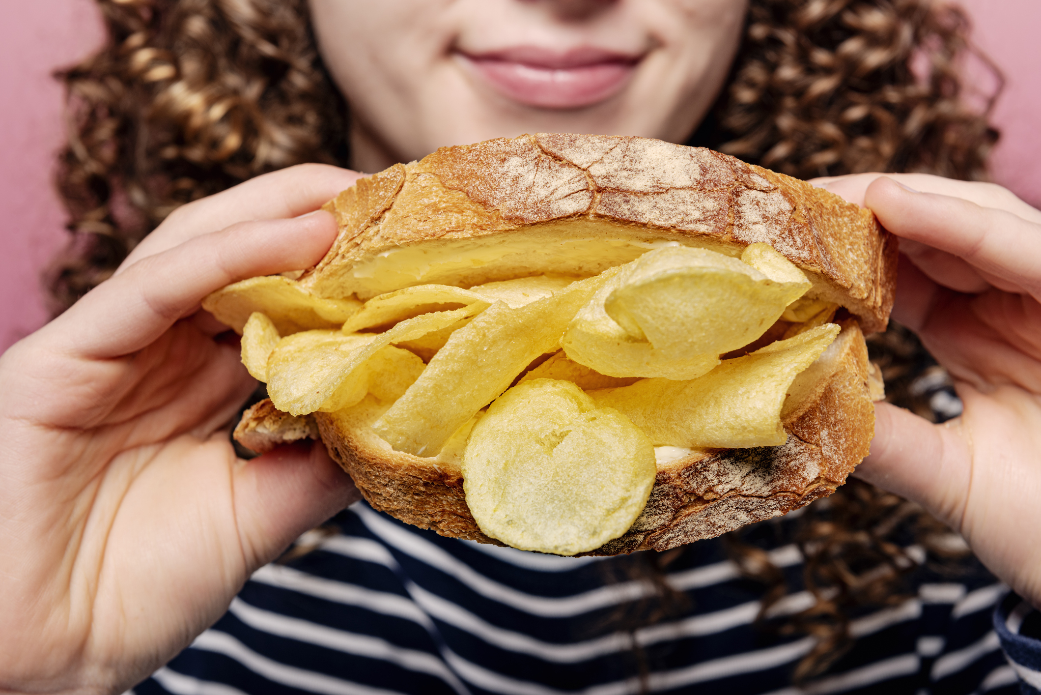Person holding and about to eat a sandwich filled with potato chips. The person's face is partially visible, showing a smile