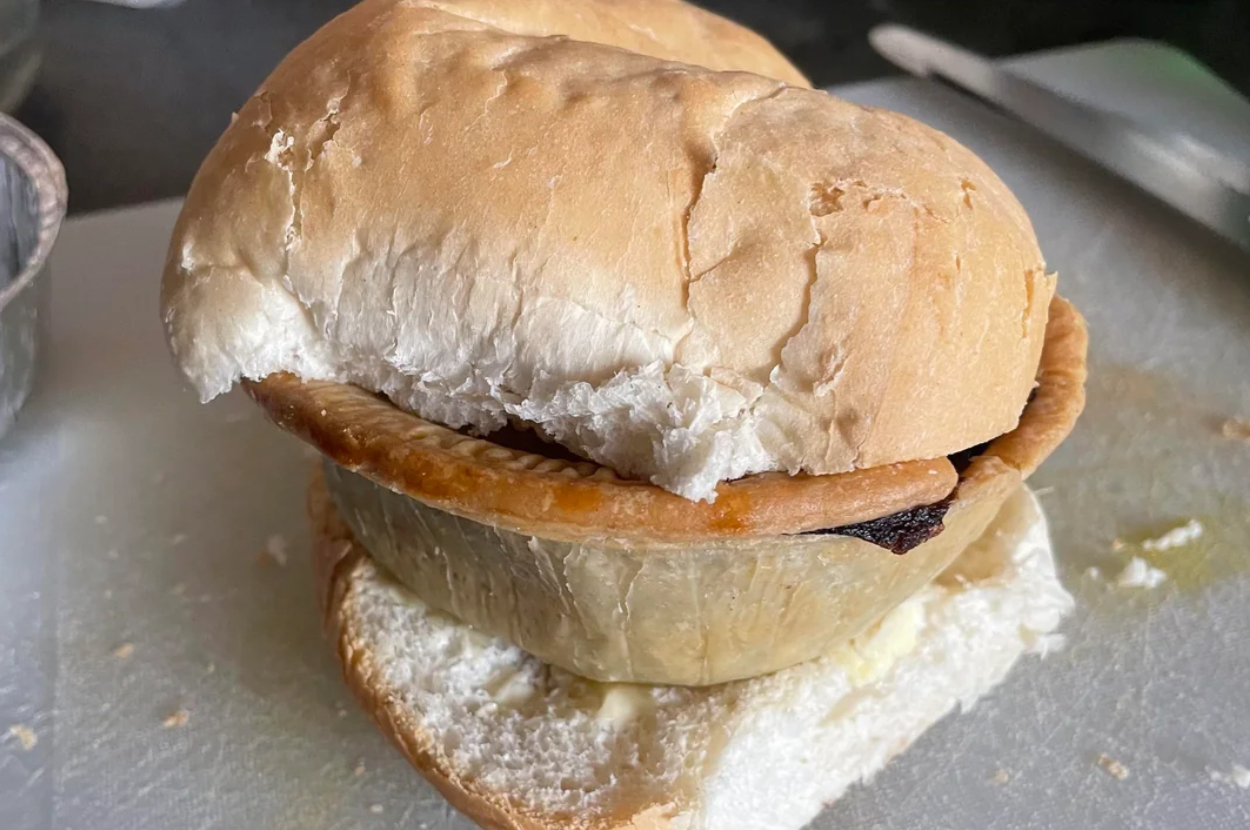 A meat pie inside a bread roll, placed on a white surface, creating a pie sandwich
