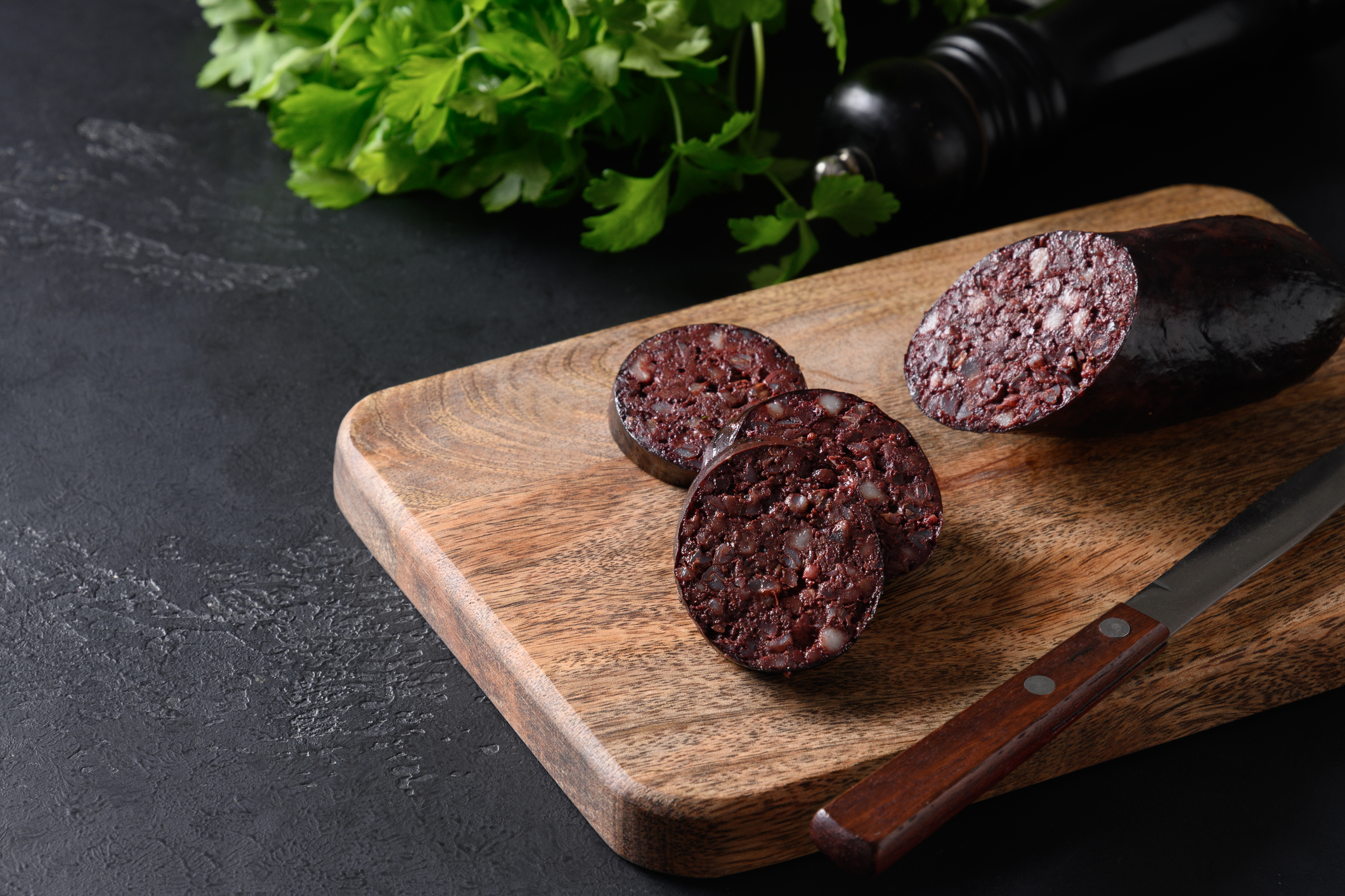 Sliced blood sausage on a wooden cutting board with a knife and fresh herbs in the background