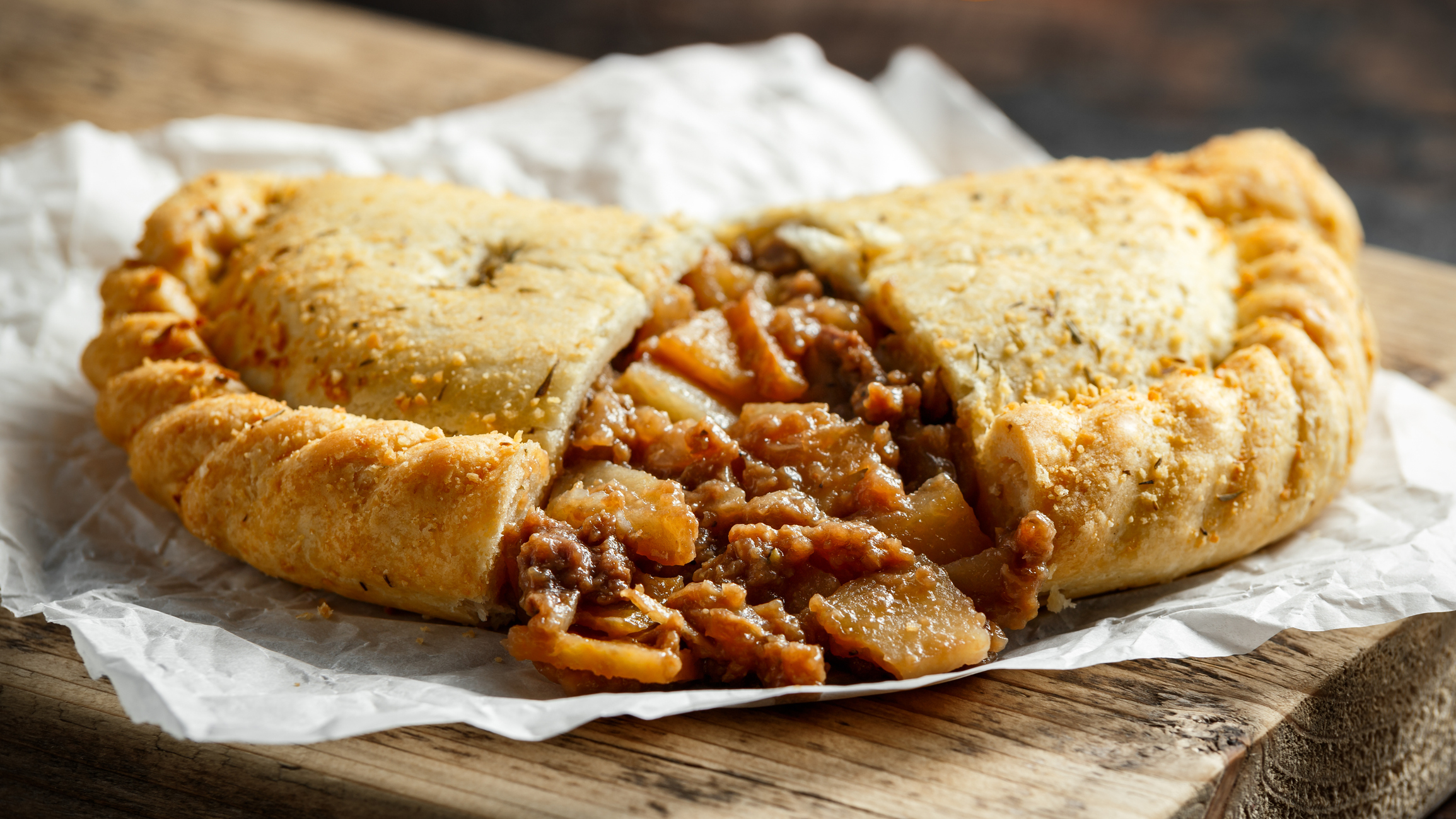 A close-up view of a baked pastry filled with spiced apple and meat filling, displayed on parchment paper over a wooden surface