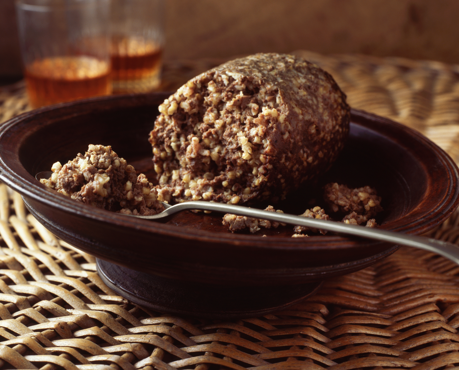 A serving of haggis on a dark wooden plate with a fork next to it, placed on a woven mat with two glasses in the background