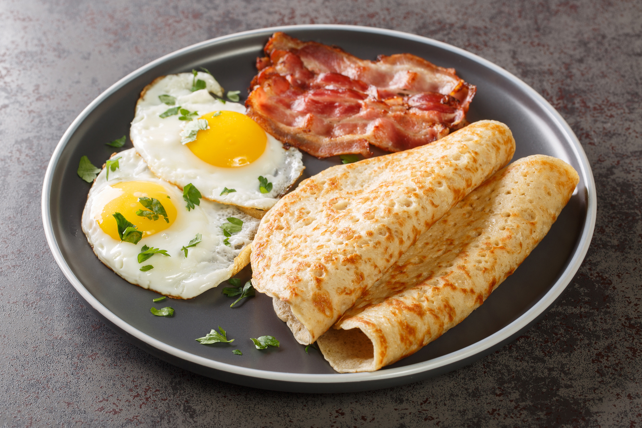 Plate with two fried eggs, bacon strips, and two rolled pancakes garnished with chopped herbs
