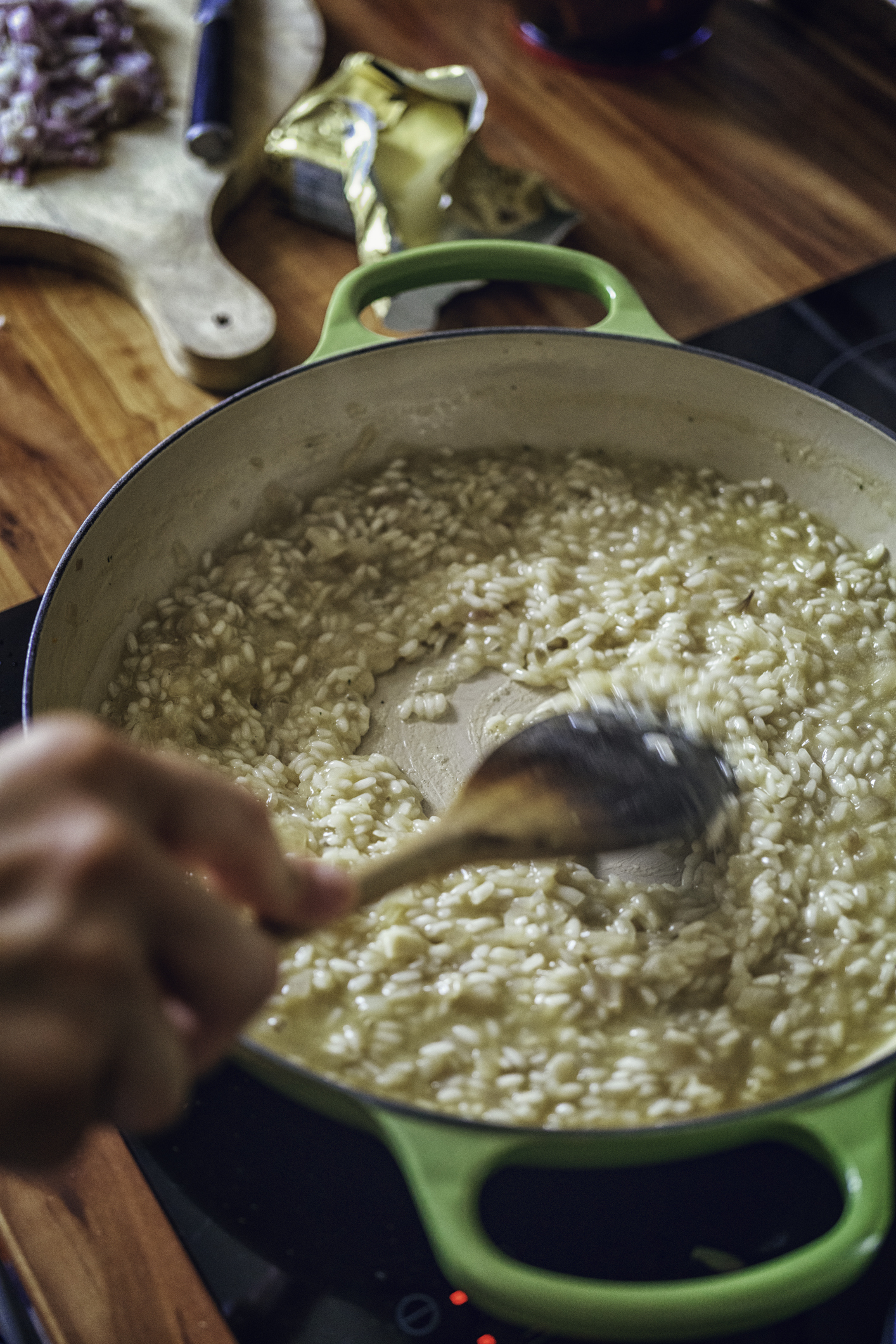 Person stirring risotto in a pot on a stovetop with a wooden spoon. Garlic cloves and ingredients are on a cutting board in the background