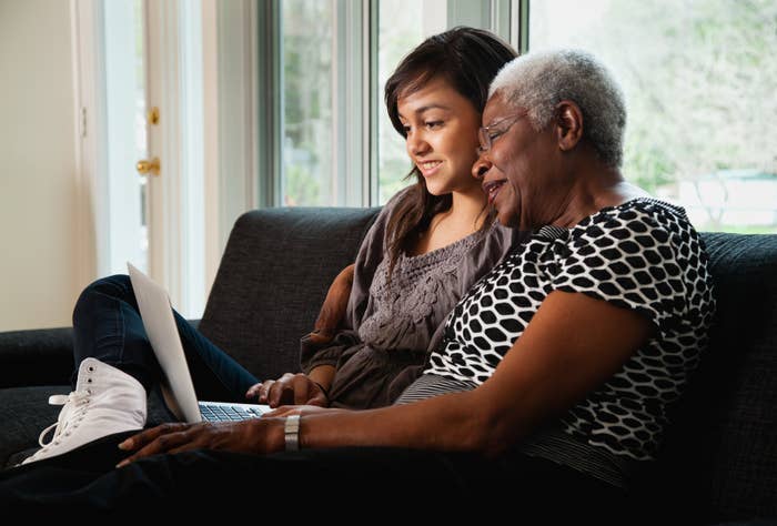A young woman and an older woman sit on a couch, smiling and looking at a laptop together