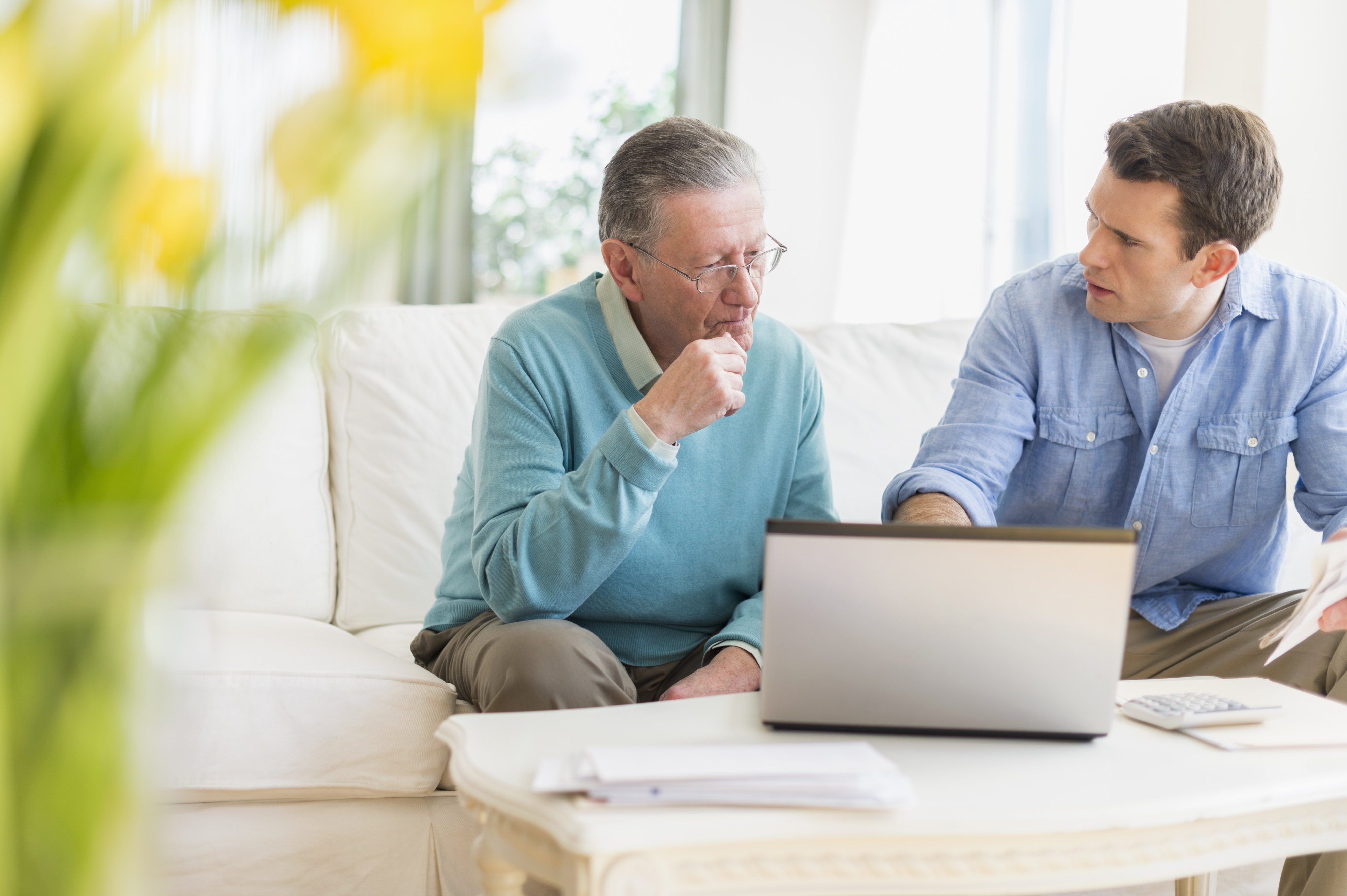 An older man and a younger man are sitting on a couch, discussing something on a laptop