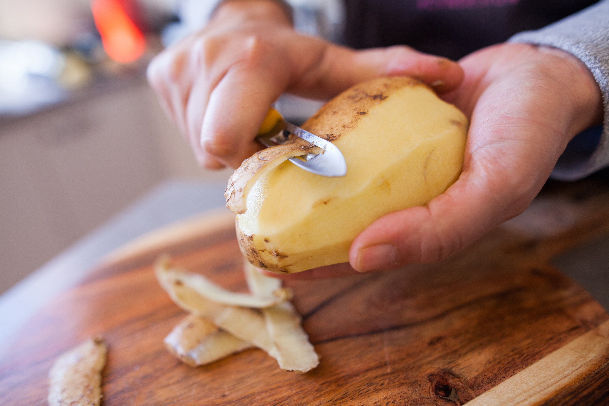 Person peeling a potato with a peeler over a wooden cutting board