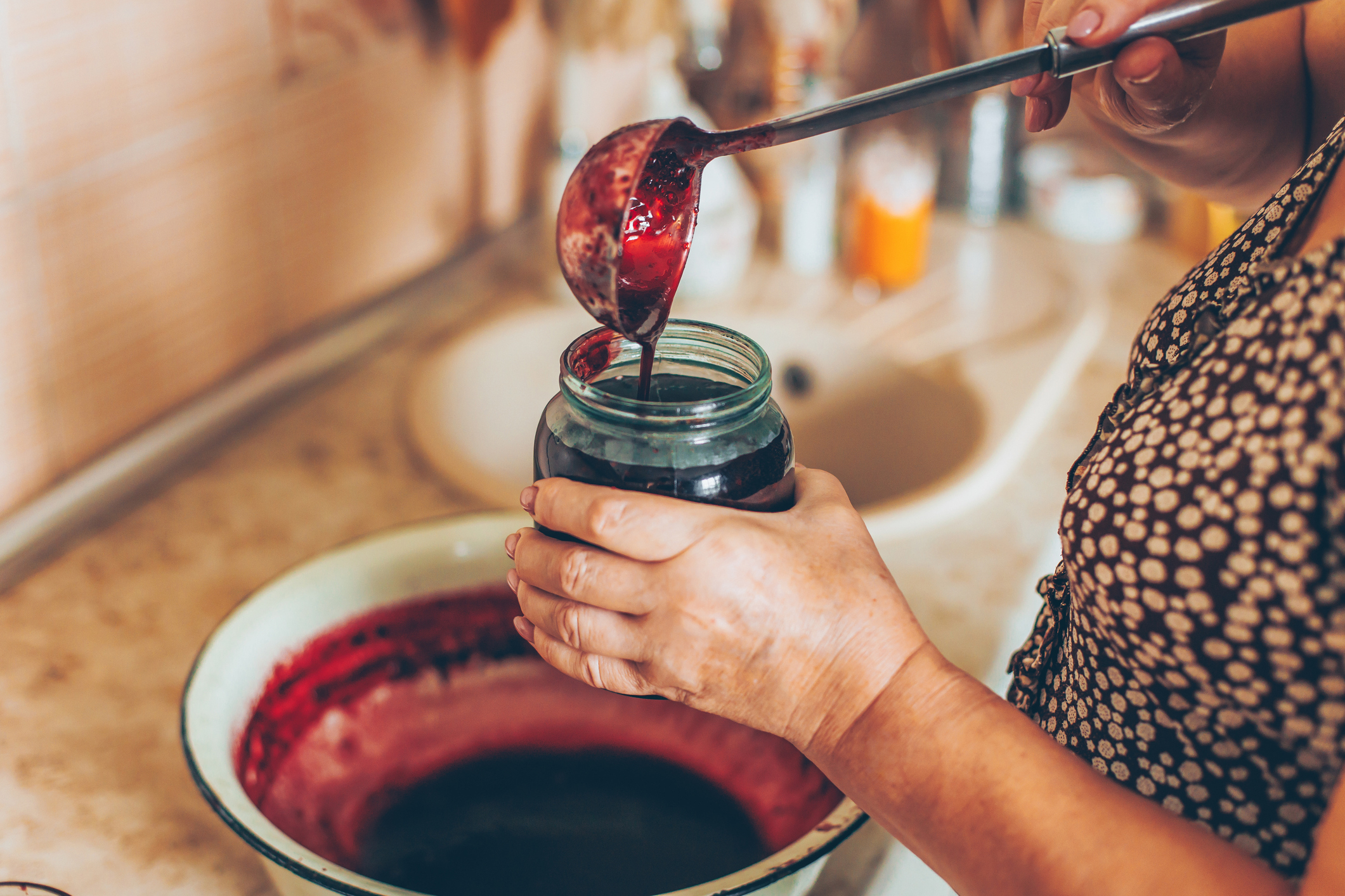 A person is ladling homemade jam into a glass jar in a kitchen setting