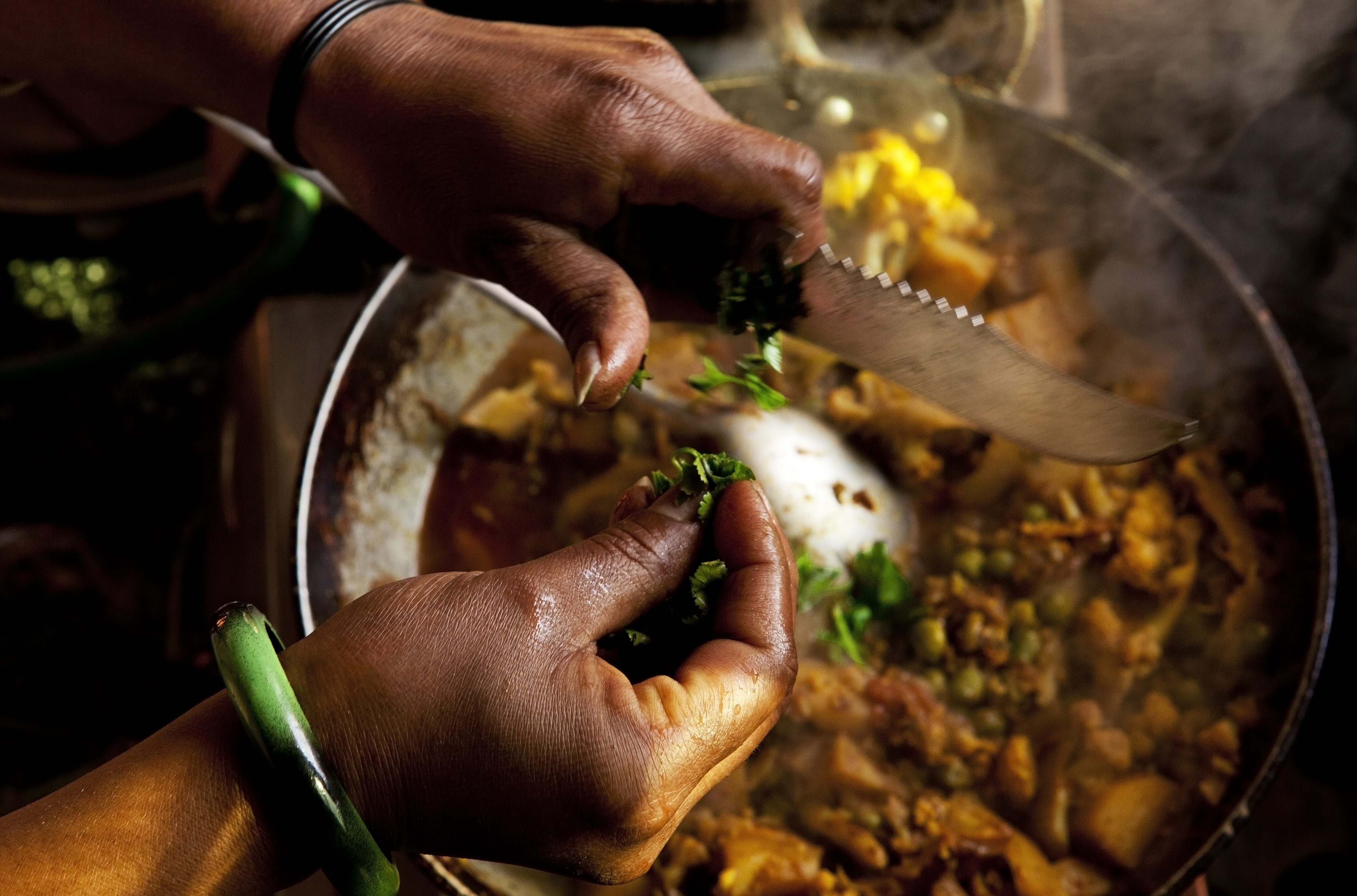 Close-up of hands preparing a dish by chopping herbs over a pan with various ingredients cooking. One wrist has a green bangle