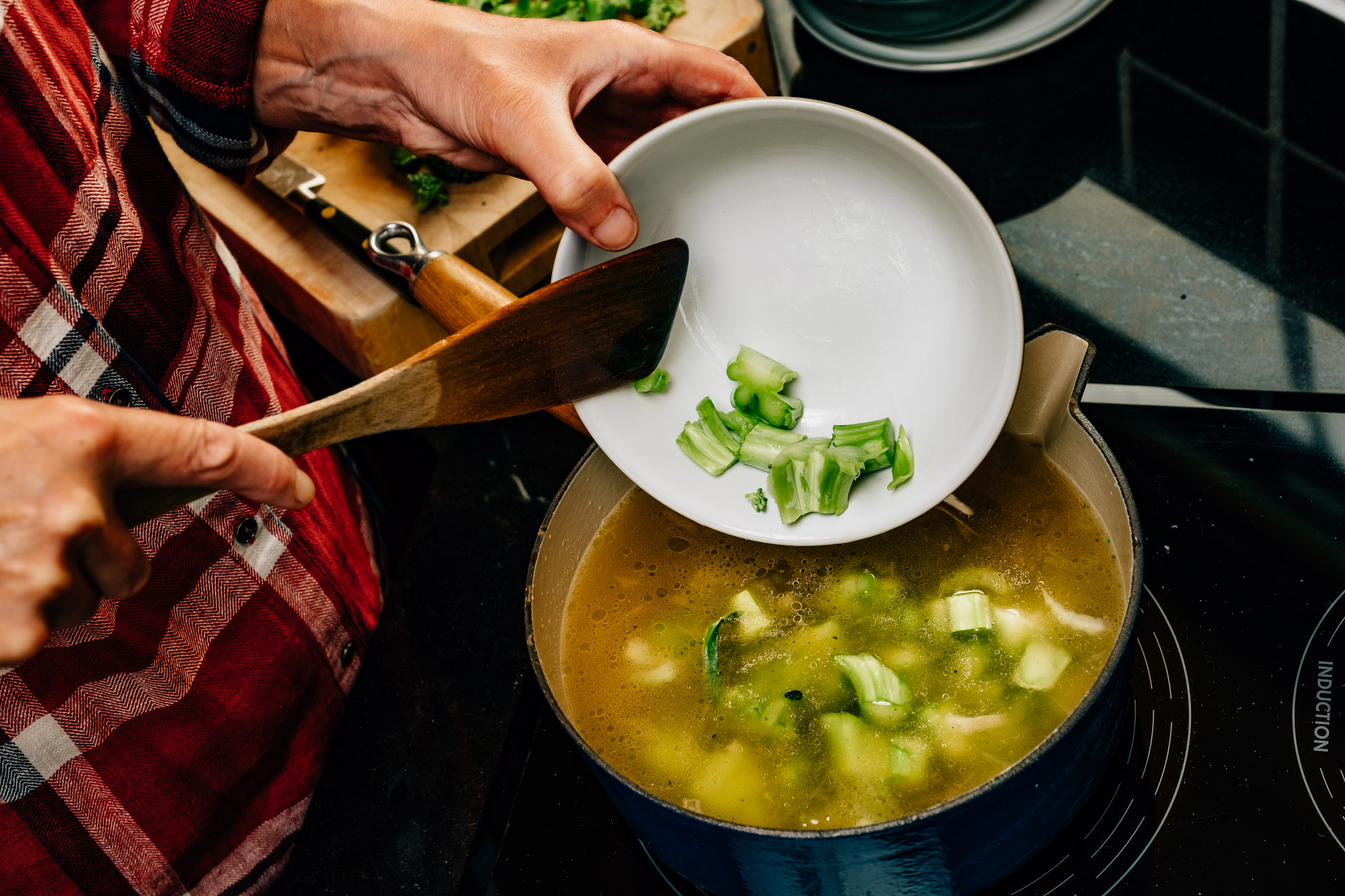 A person is adding chopped celery from a white bowl into a pot of soup simmering on a stovetop