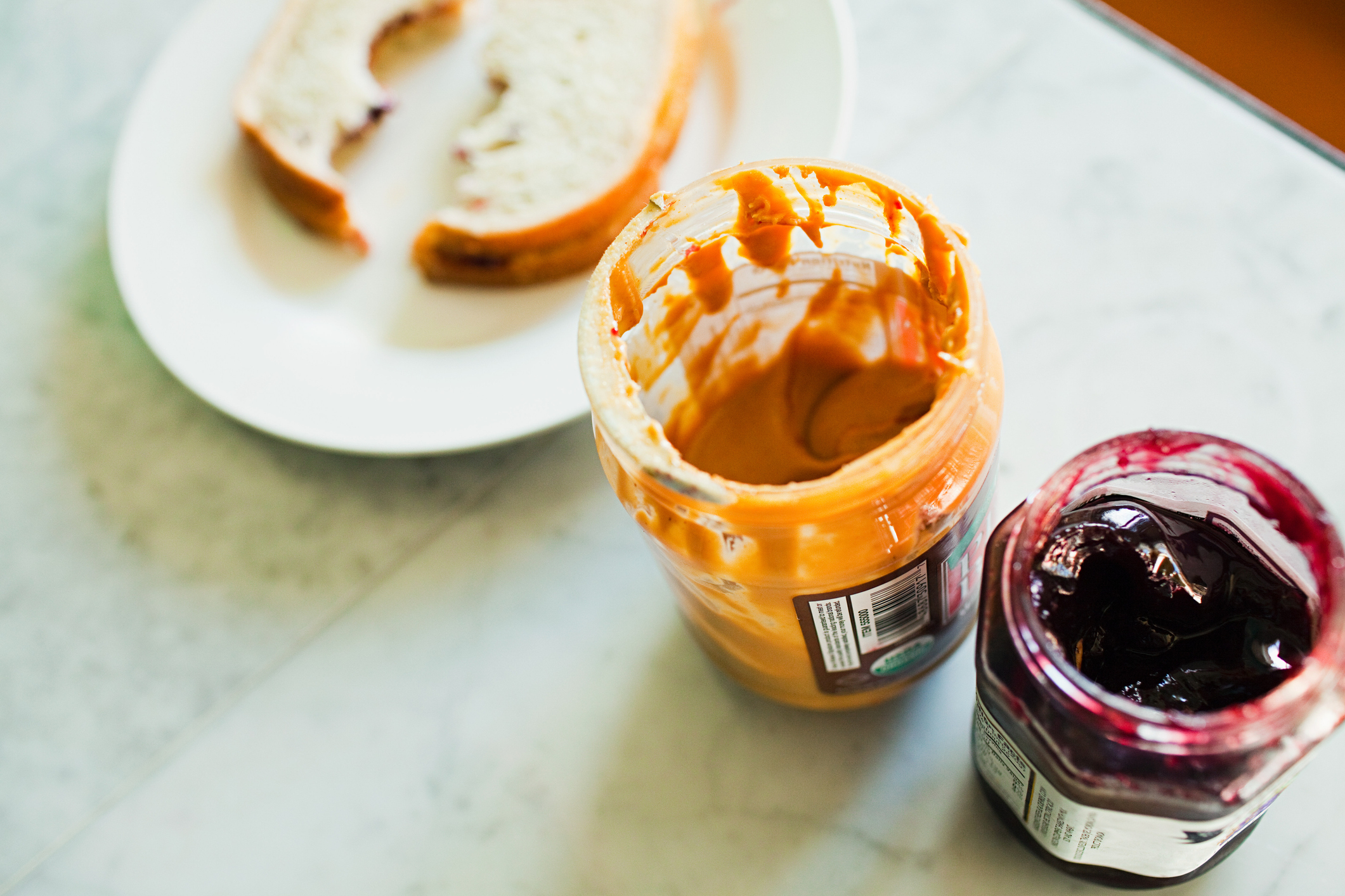 A jar of peanut butter and a jar of jelly sit open on a marble countertop. In the background, there's a plate with two slices of bread