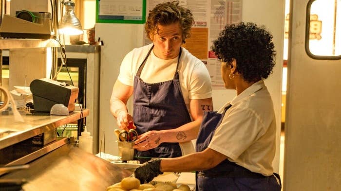 Chef Carmen &quot;Carmy&quot; Berzatto and Sydney Adamu from &quot;The Bear&quot; prepare food in a commercial kitchen. Carmy is in a white shirt and blue apron; Sydney is in a white shirt and blue apron