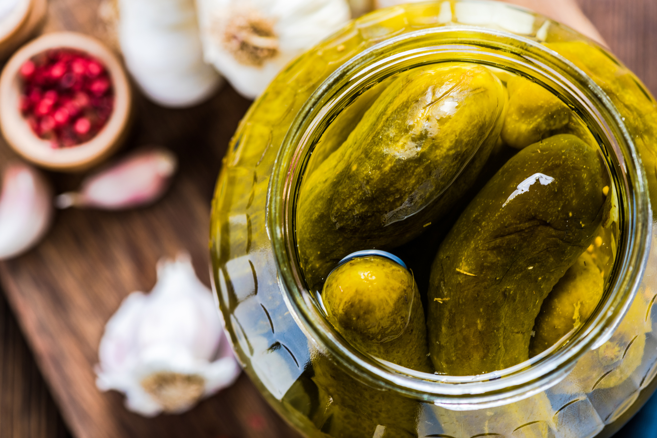 An open jar filled with pickles on a wooden table, surrounded by garlic cloves and a small bowl of spices