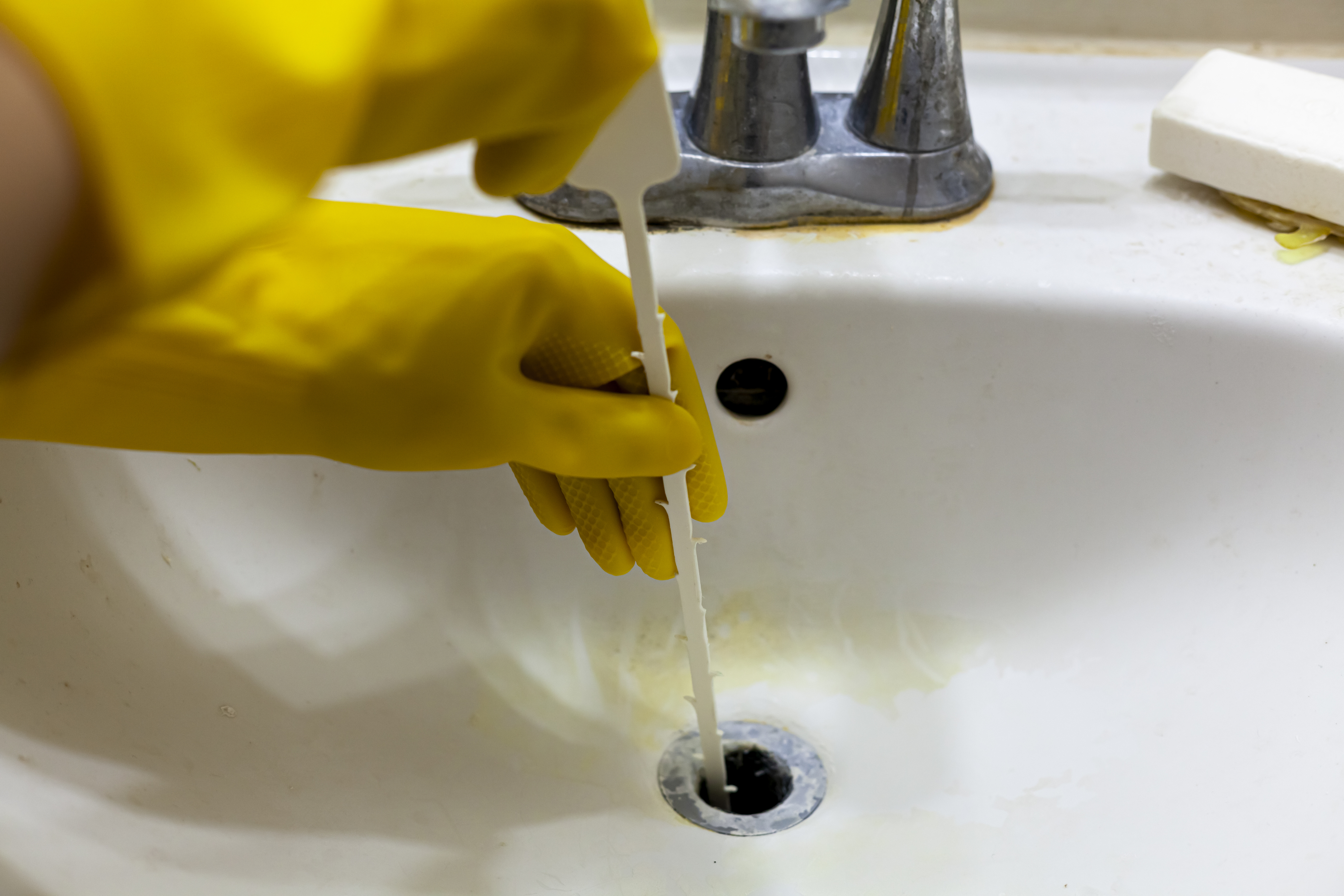 A person wearing yellow gloves uses a plastic drain snake to unclog a bathroom sink