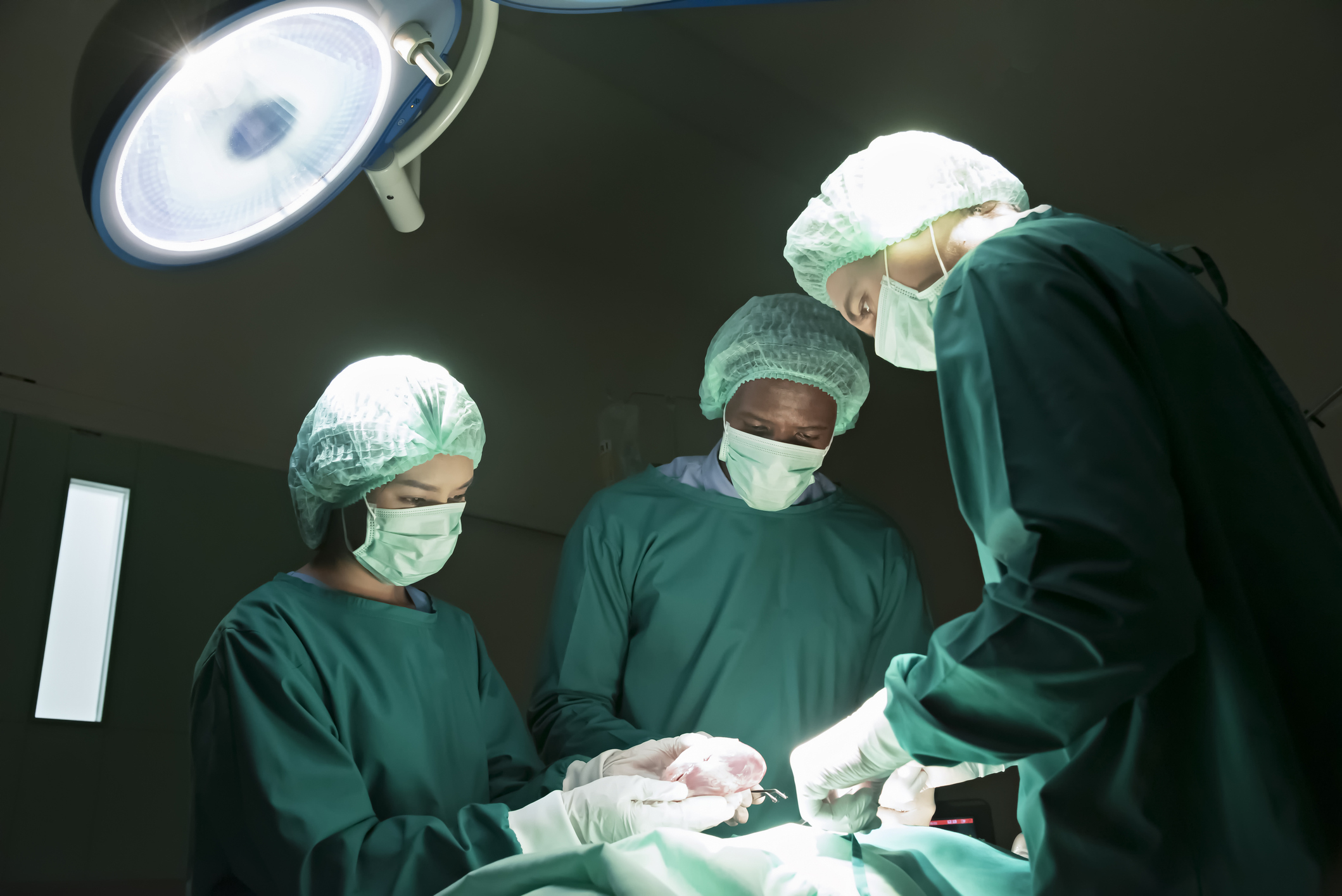 Three surgeons in operating room performing surgery, all wearing surgical gowns, gloves, masks, and caps. Focused and serious