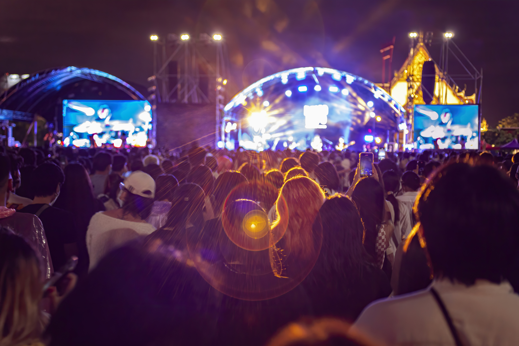 A large crowd watches a nighttime outdoor concert with bright stage lights and large video screens displaying the performers