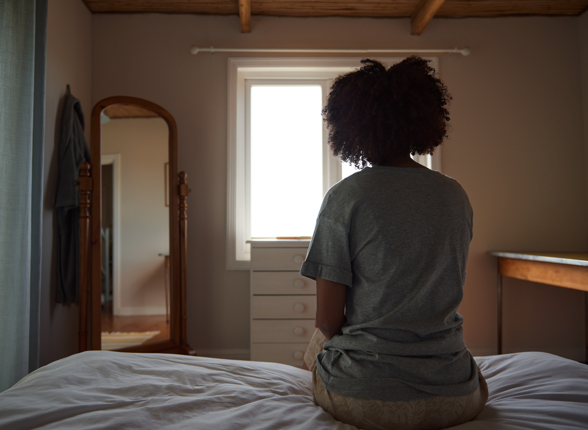 Person with curly hair sits on a bed facing a window, back to the camera, in a simple, tidy bedroom with a dresser and mirror