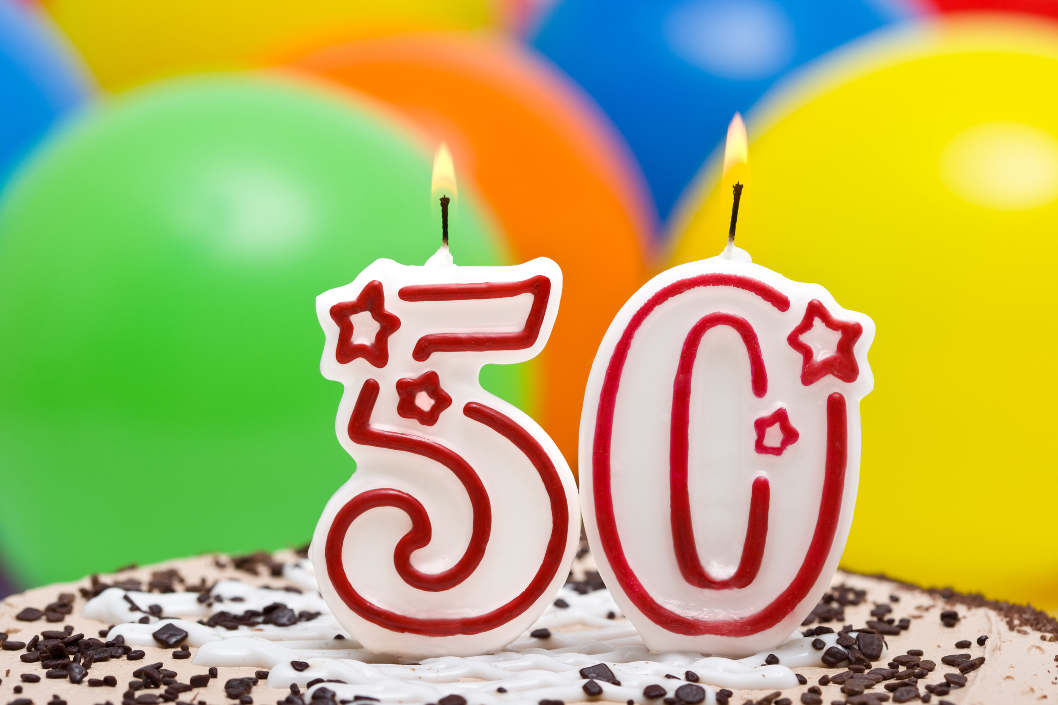 Birthday cake with lit candles shaped as the number 50, against a background of colorful balloons