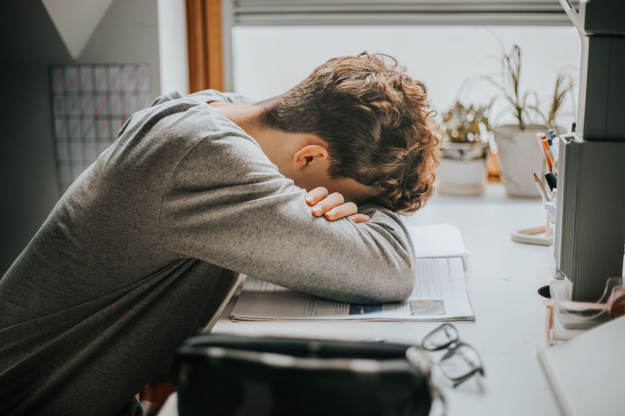 A person with their head resting on their arms at a desk, looking stressed or tired. The desk has various stationery items and plants
