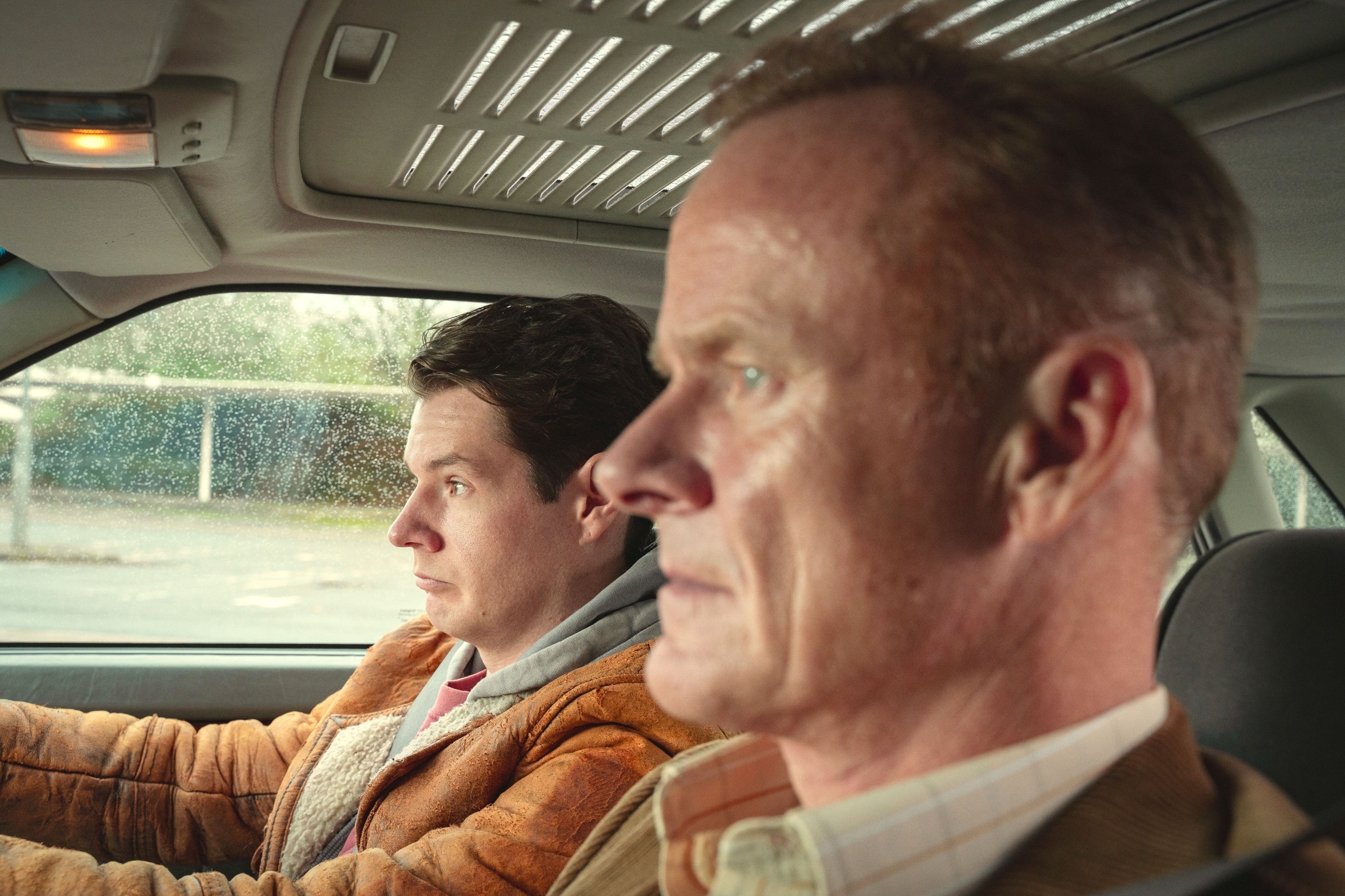 Two men, one driving and the other in the passenger seat, appear focused and serious inside a car