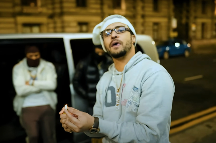 A man in a hoodie and cap is lighting an object in his hand, standing in front of a van with another person inside on a city street at night
