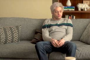 Pete Davidson sitting on a couch, wearing a light sweater with stripes and jeans, looking thoughtful