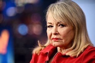 Roseanne Barr, wearing a jacket, looking at the camera with a serious expression, appears in an interview setting