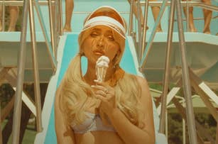 Sabrina Carpenter, holding an ice cream cone, wears a checkered bikini and a visor, standing in front of a pool slide in the Espresso music video