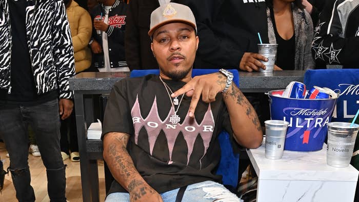 Bow Wow poses for a photo while seated at an event. He wears a cap, graphic t-shirt, and ripped jeans, gesturing with his hand. Other people are in the background