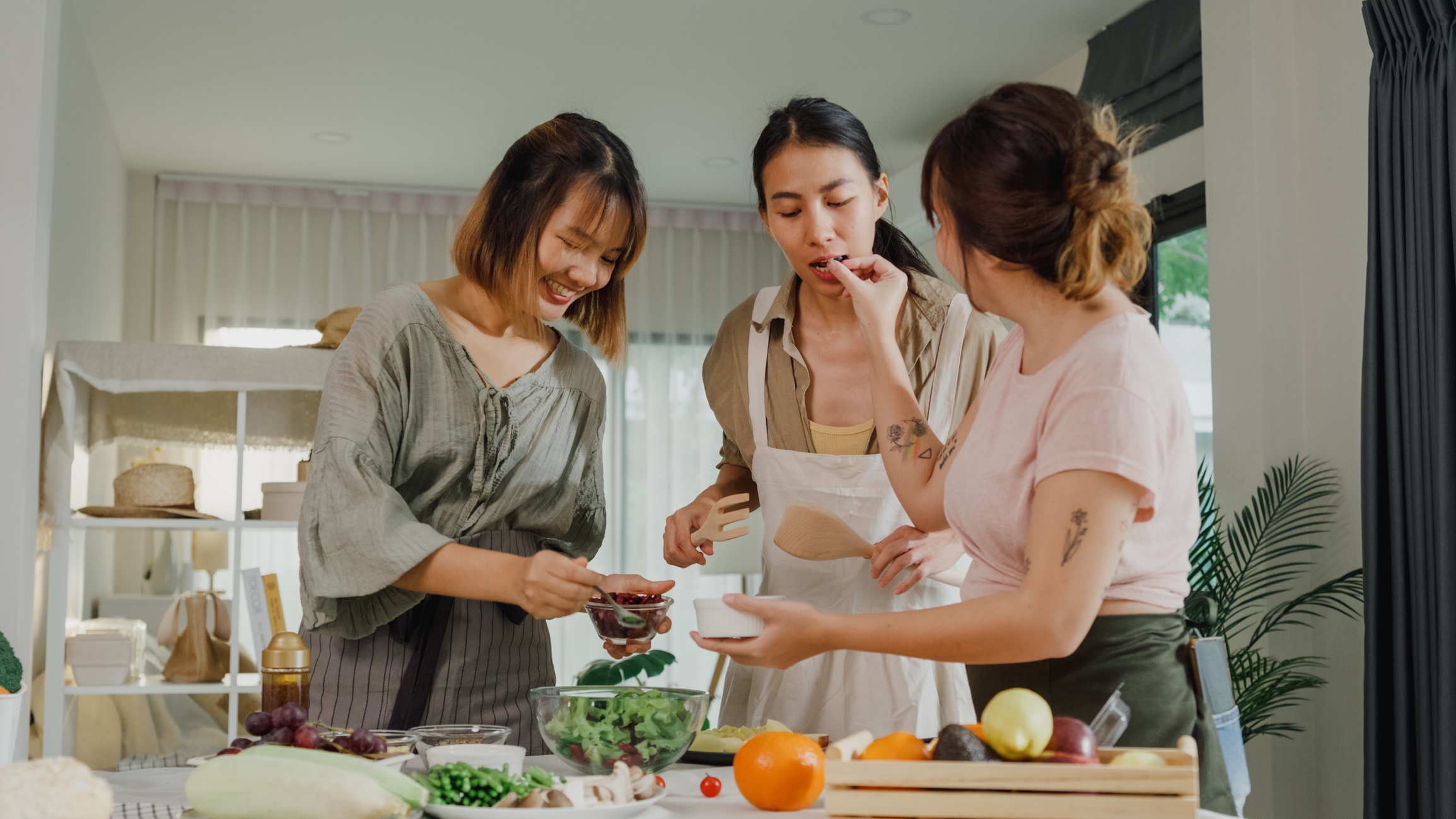 Three women, names unknown, preparing vegetables and fruit together in a kitchen, smiling and tasting food