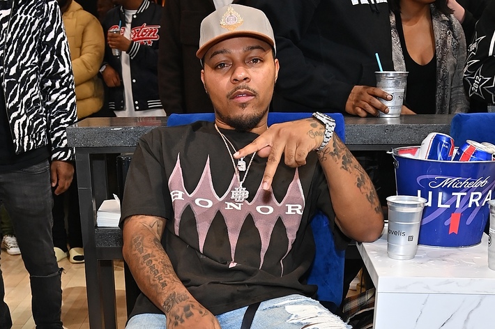 Bow Wow sits casually at an event, wearing a cap and a black Honor T-shirt with ripped jeans, making a hand gesture. Several people are in the background