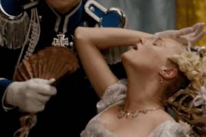 Character fainting in a historical style film scene. The character is dressed in period clothing with elaborate accessories, being fanned by another individual