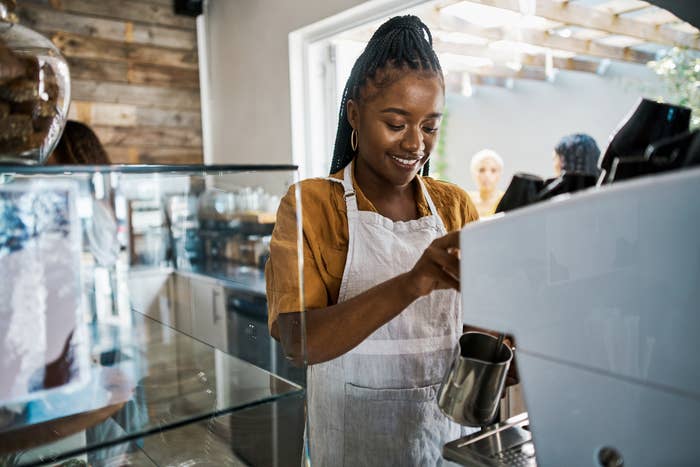 Woman in an apron smiling while preparing a coffee drink behind the counter of a café. Other patrons and café interiors are visible in the background