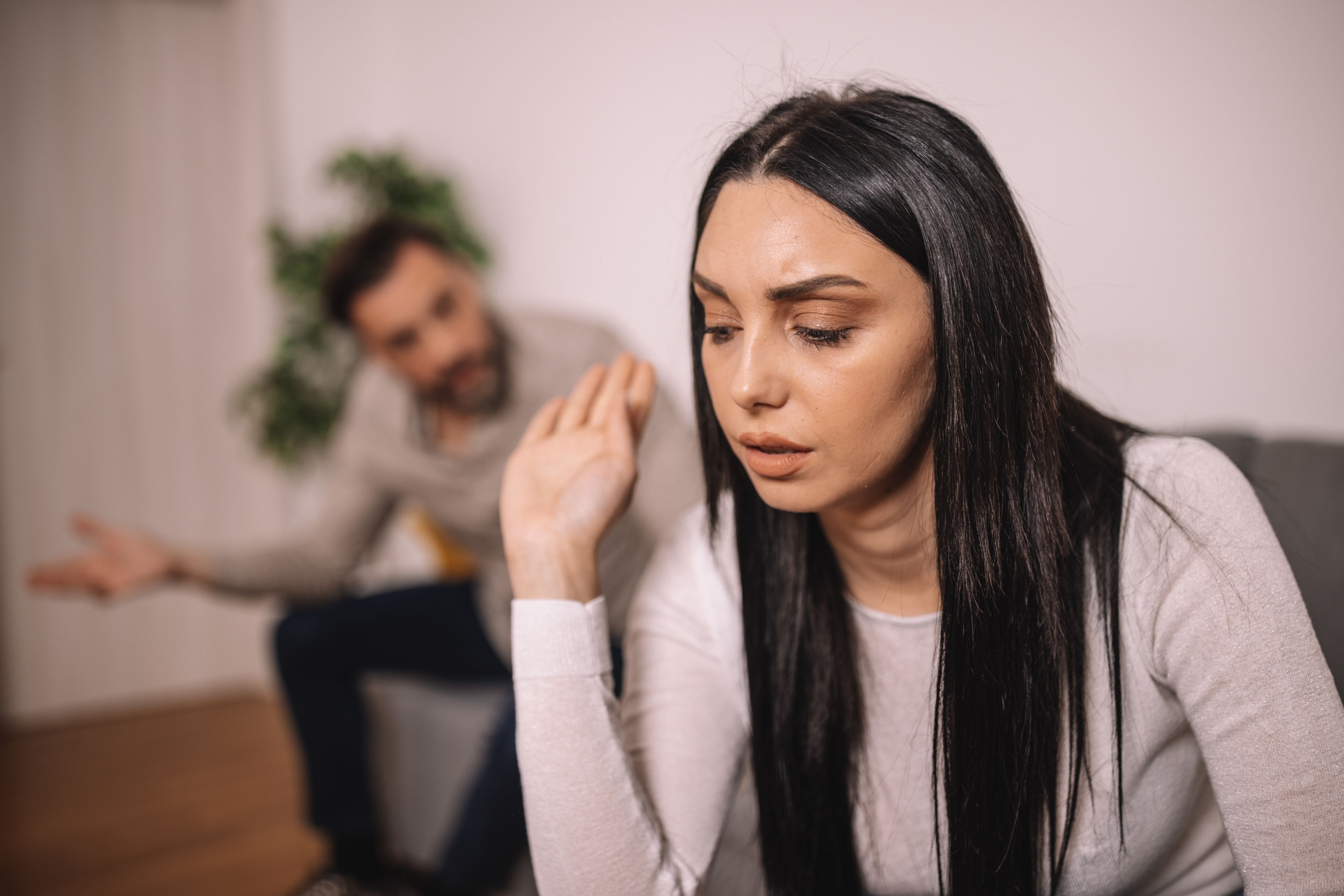 A woman with long dark hair looks upset and gestures dismissively while a man in the background appears to be arguing or talking to her