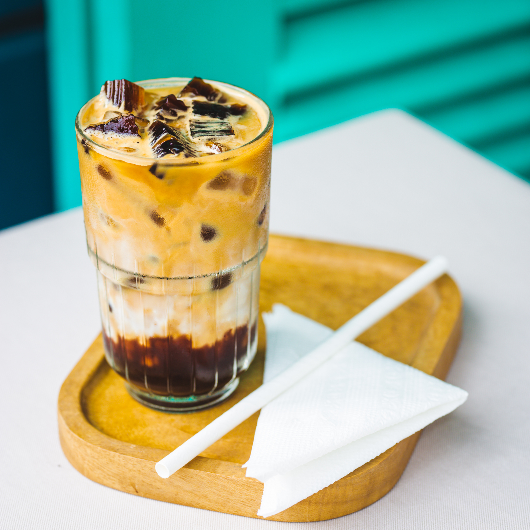 Glass of iced coffee with layered cream and chocolate chunks, served on a wooden tray with a straw and napkin