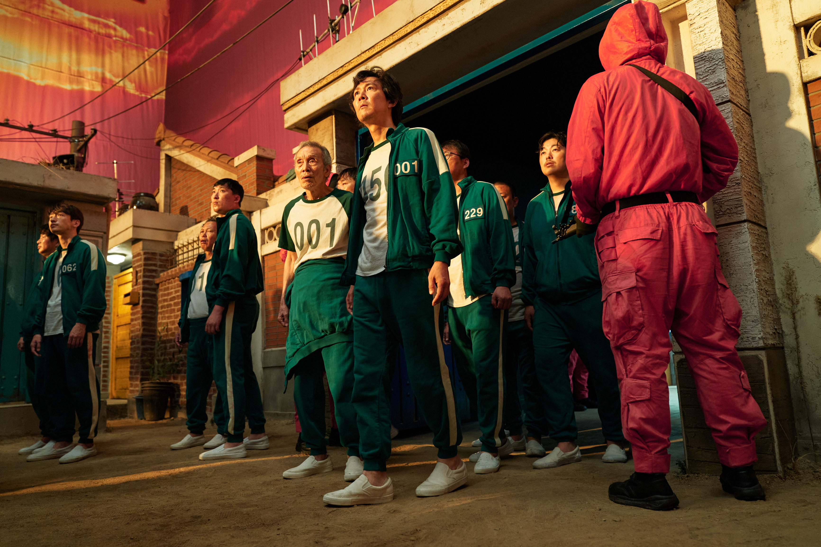 "Squid Game cast in green tracksuits with numbers, standing on set with a person in a pink jumpsuit and black mask."