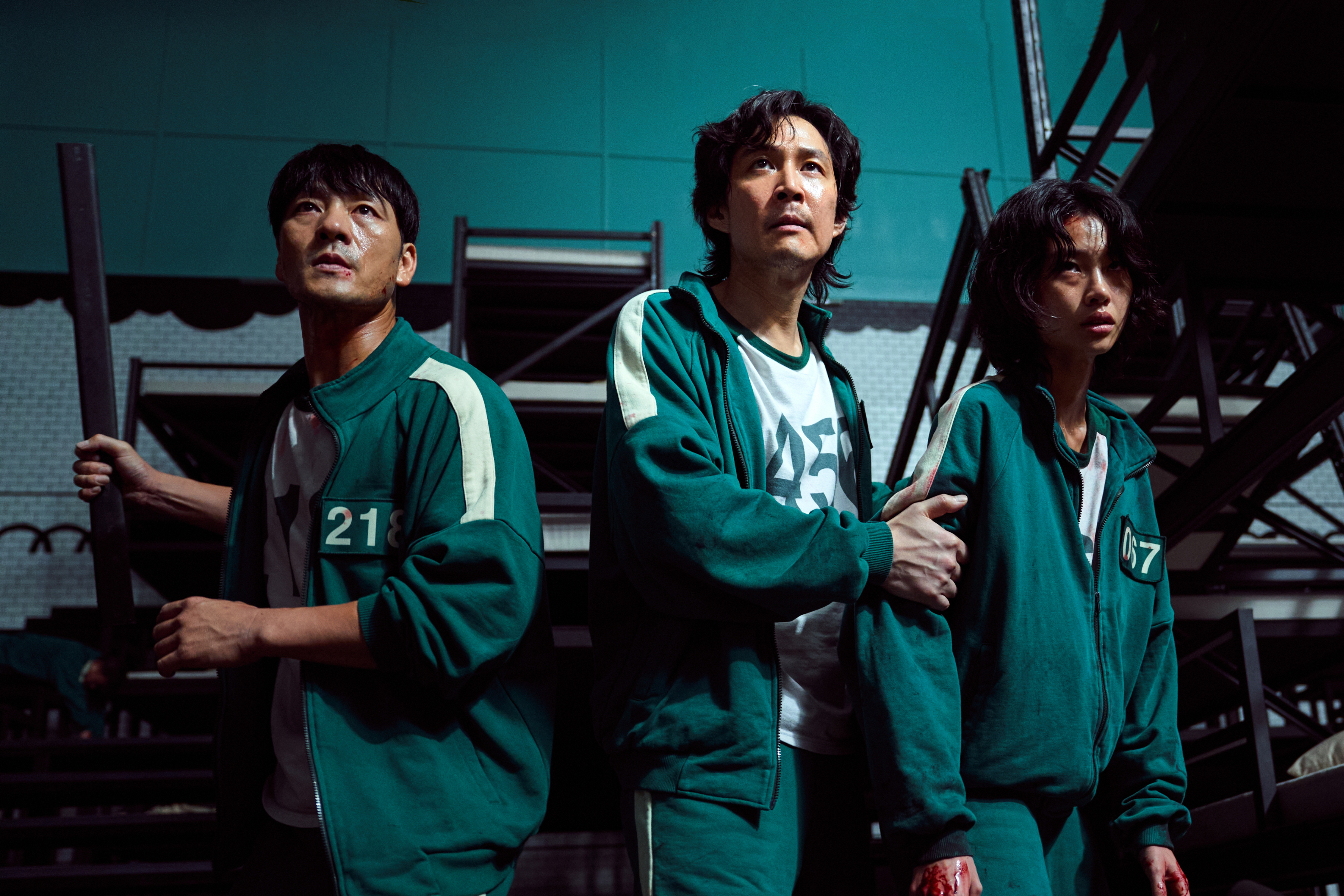 Three actors from Squid Game, wearing numbered tracksuits, stand in a tense scene. The one on the right is holding his bleeding hand