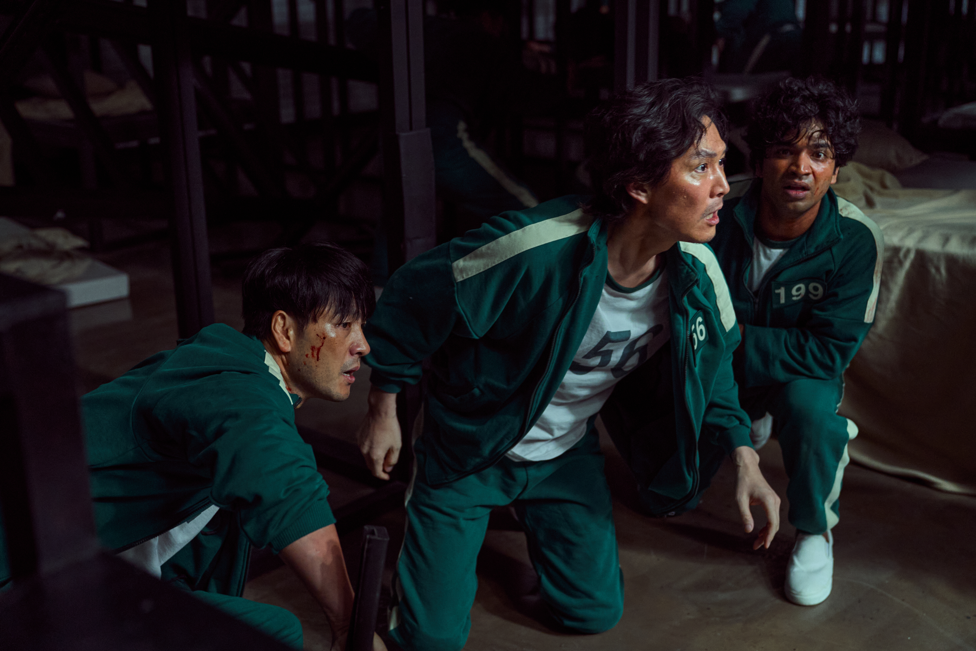 Three actors from "Squid Game" in green tracksuits with numbers 456 and 199 appear tense inside a dimly lit dormitory setting