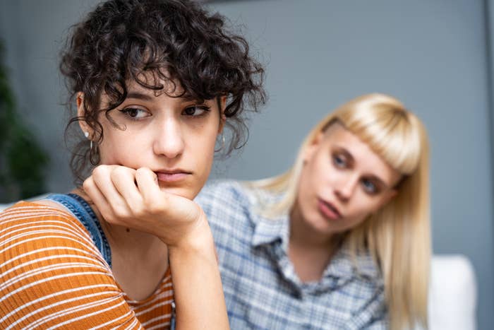 Woman in foreground with curly hair looks away sadly, resting her chin on her hand. In the background, another woman with blonde hair looks at her with concern