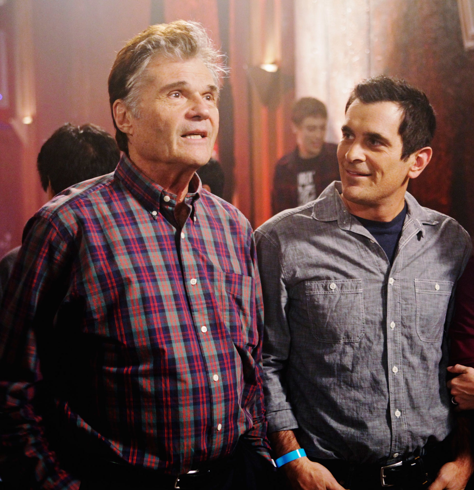 Fred Willard in a plaid shirt, Ty Burrell in a casual shirt, and Julie Bowen in a dress stand together, smiling, in a social setting
