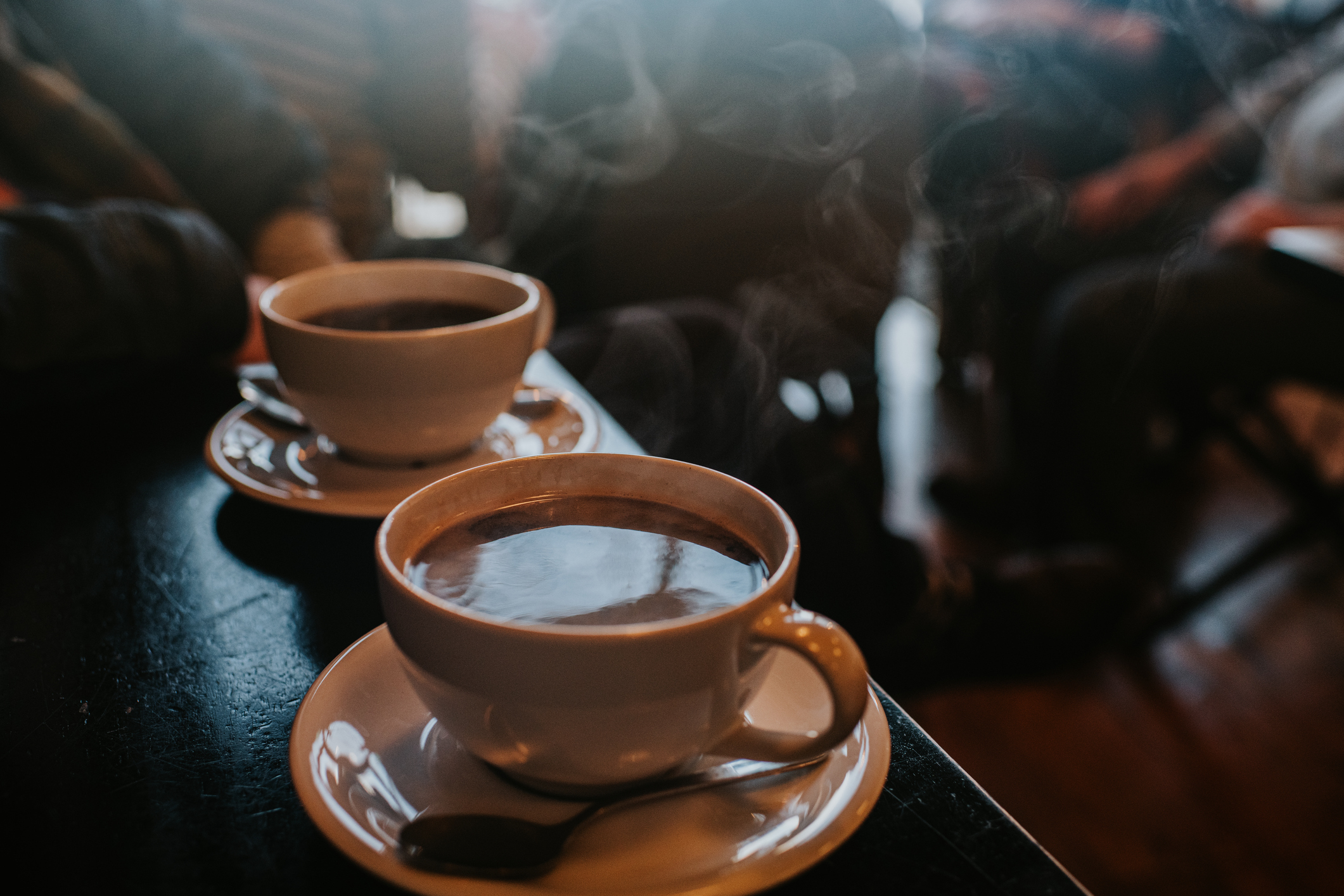Two steaming cups of coffee on a table in a coffee shop, with blurred people sitting in the background