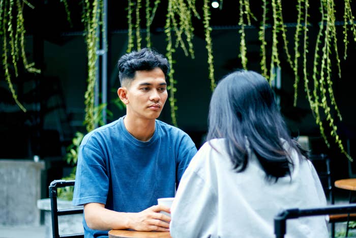 A person in a blue shirt sits across from a person in a white shirt at an outdoor café. Both are engaged in conversation