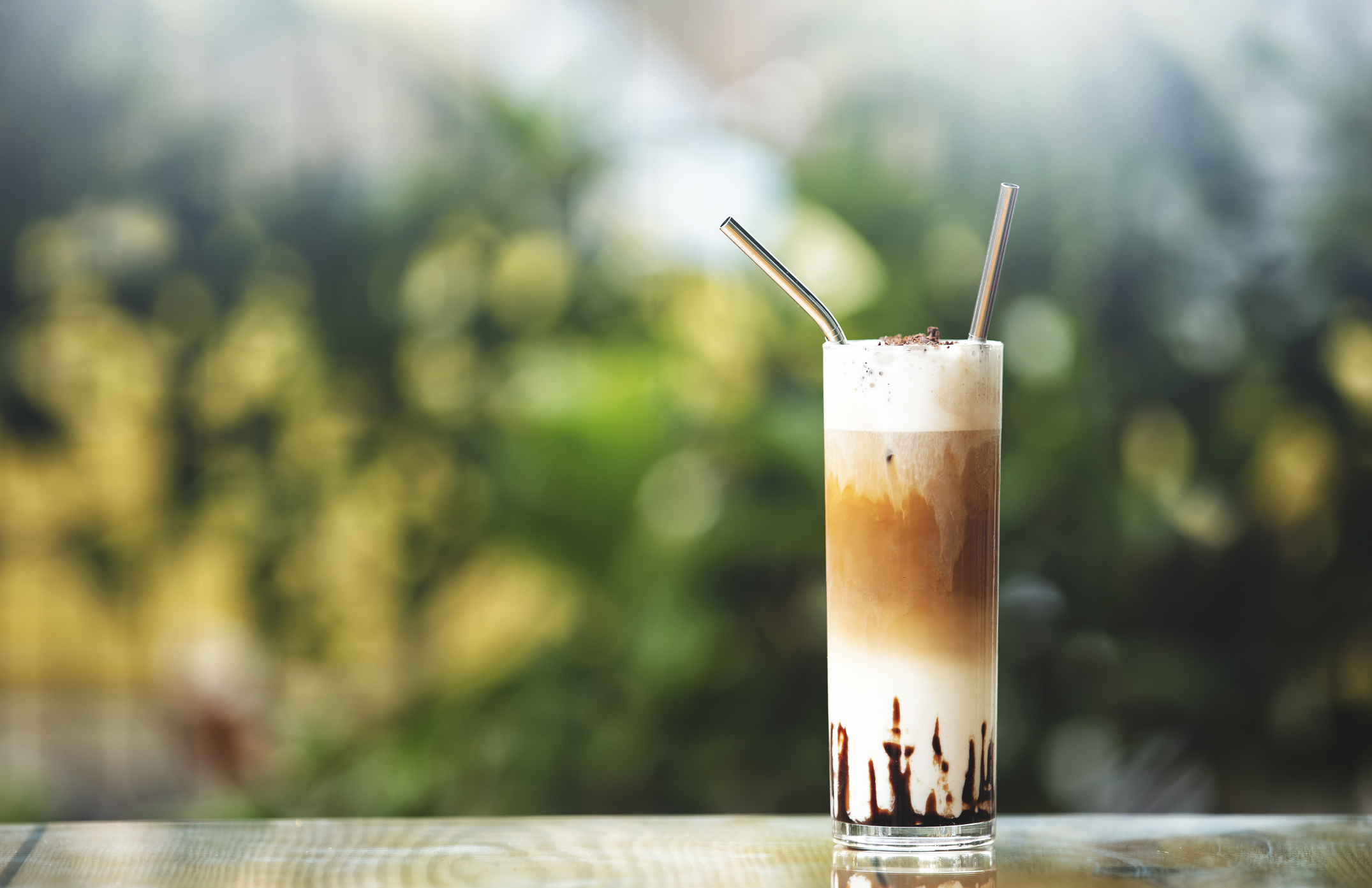 A tall glass of iced coffee is topped with foam and served with two metal straws, placed on a wooden table with a blurred natural background