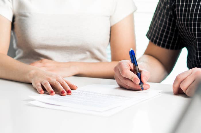 Two people, one in a short-sleeve top and the other in a checkered short-sleeve shirt, reviewing and signing a document on a table
