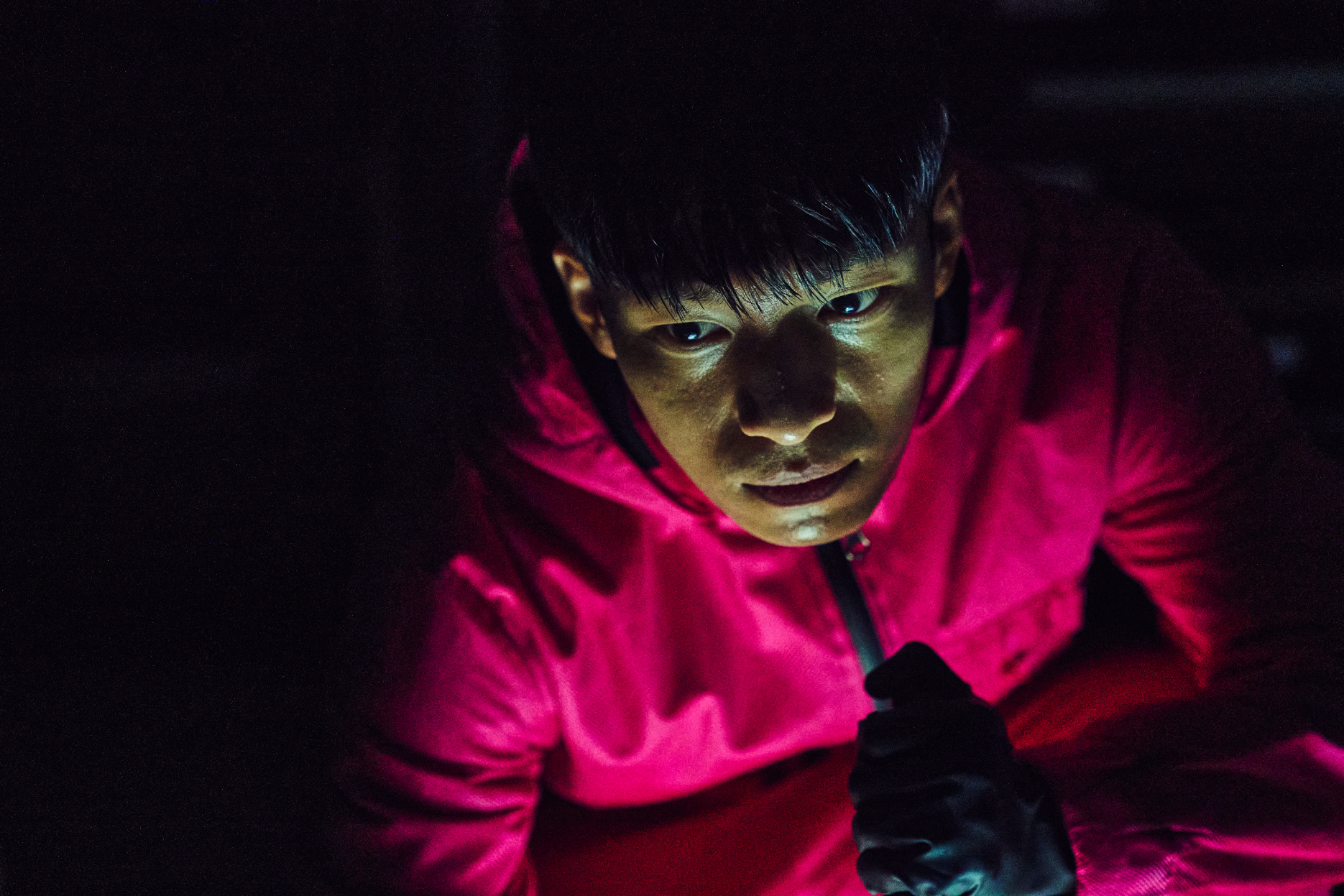 Woo-sik Choi, wearing a hooded outfit, crouches in a dimly lit scene, holding a flashlight up to his face